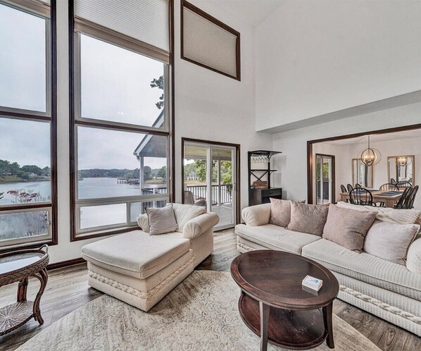 Ein geräumiges Wohnzimmer mit großen Fenstern und Blick auf ein Gewässer, eine gemütliche Sitzecke mit Sofa und Hocker sowie ein Essbereich mit einer Chandelüster.