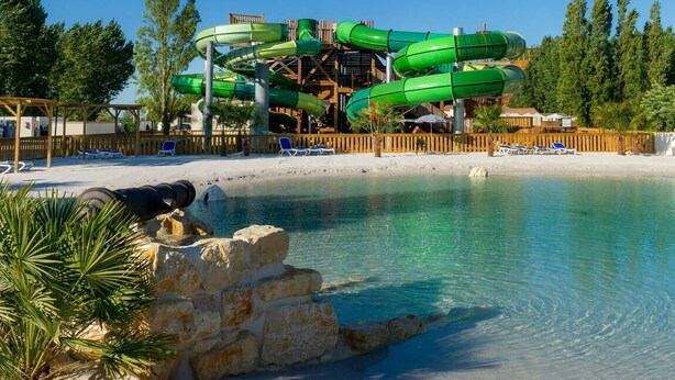 Ein Wasserpark mit mehreren grünen Rutschen, einem Sandstrand und klarem Wasser.