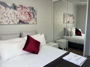 A hotel room with a large bed, white bedding, a red pillow, a small bedside table, a mirror, and a floral artwork on the wall.