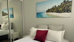 En pænt redt seng med hvide og røde puder, et hvidt natbord og et sammenfoldet hvidt håndklæde på gulvet.