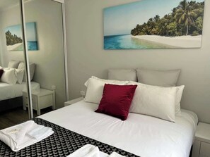 A neatly made bed with white and red pillows, a white bedside table, and a folded white towel on the floor. - Lambert's Beach Awaits with Wifi (Slade Point)