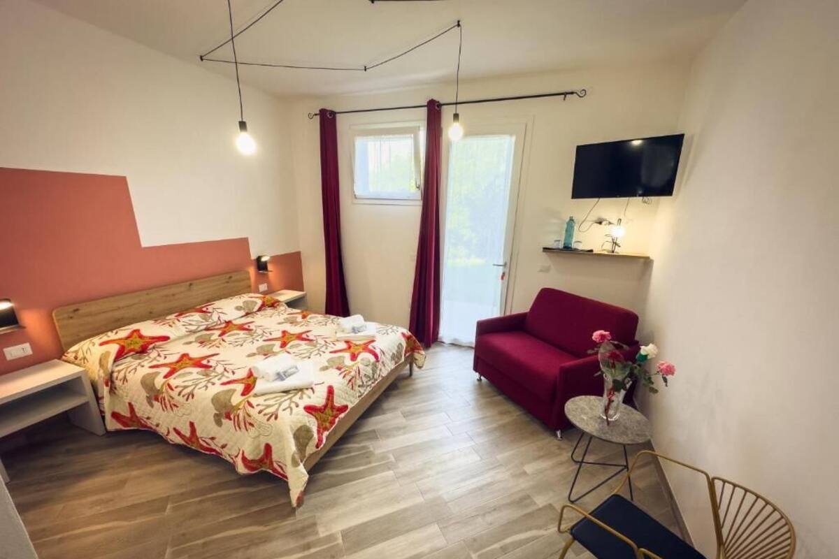 A bedroom with a bed, a red sofa, a small round table with a vase of flowers, a TV mounted on the wall, and a window with curtains.