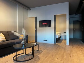 Ein modernes Wohnzimmer mit einer Couch, einem Couchtisch, einem an der Wand montierten Fernseher und einer Tür zu einem Schlafzimmer.