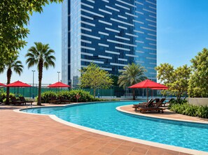 A swimming pool with lounge chairs and red umbrellas, surrounded by palm trees and a tall building in the background. - Vanilla Homes Premium 1BR in Reem Island (Abu Dhabi)