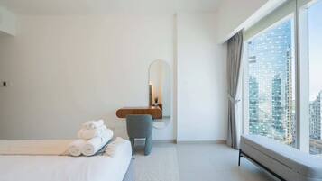 A modern hotel room with a bed, a small seating area, a mirror, and a large window with a city view.