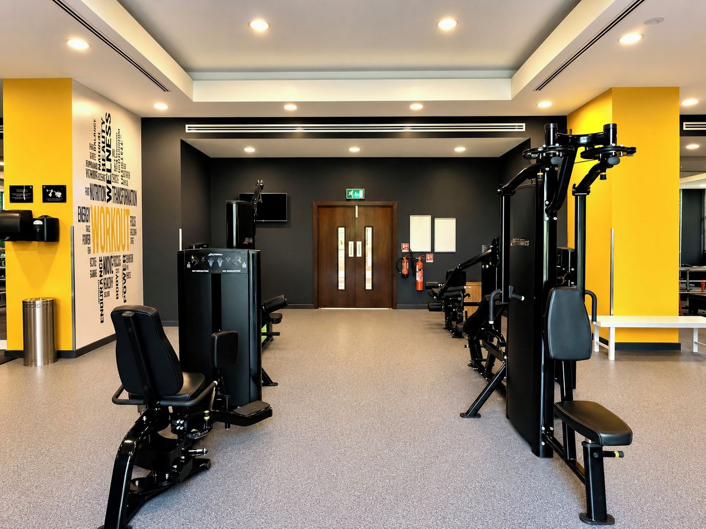 Een sportschool met fitnessapparatuur, een muur met motiverende woorden en een deur met nooduitgangsborden.