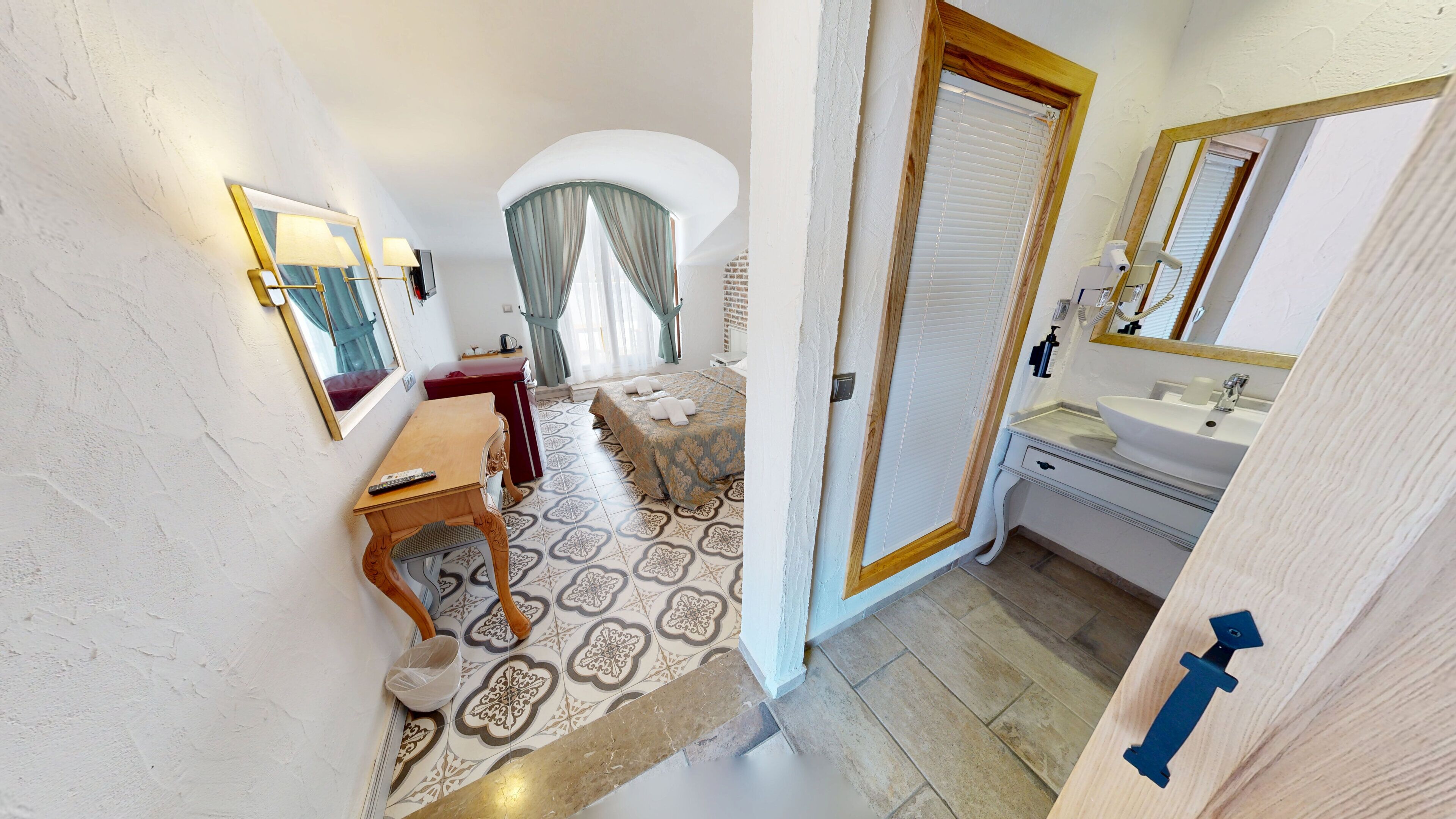 A bedroom with a bed, a desk, a chair, a mirror, and a bathroom with a sink and mirror.