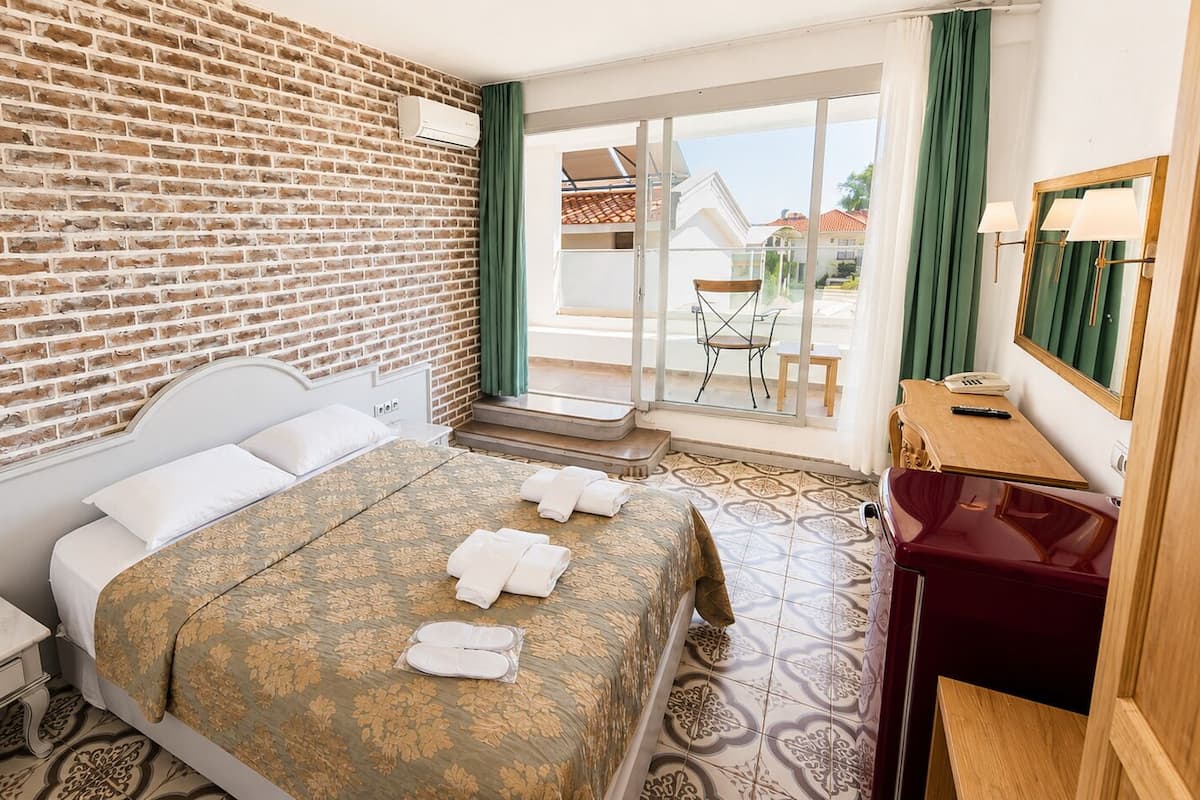 A hotel room with a brick wall, a bed with a floral bedspread, a wooden nightstand, a desk with a mirror, and a view of a balcony.