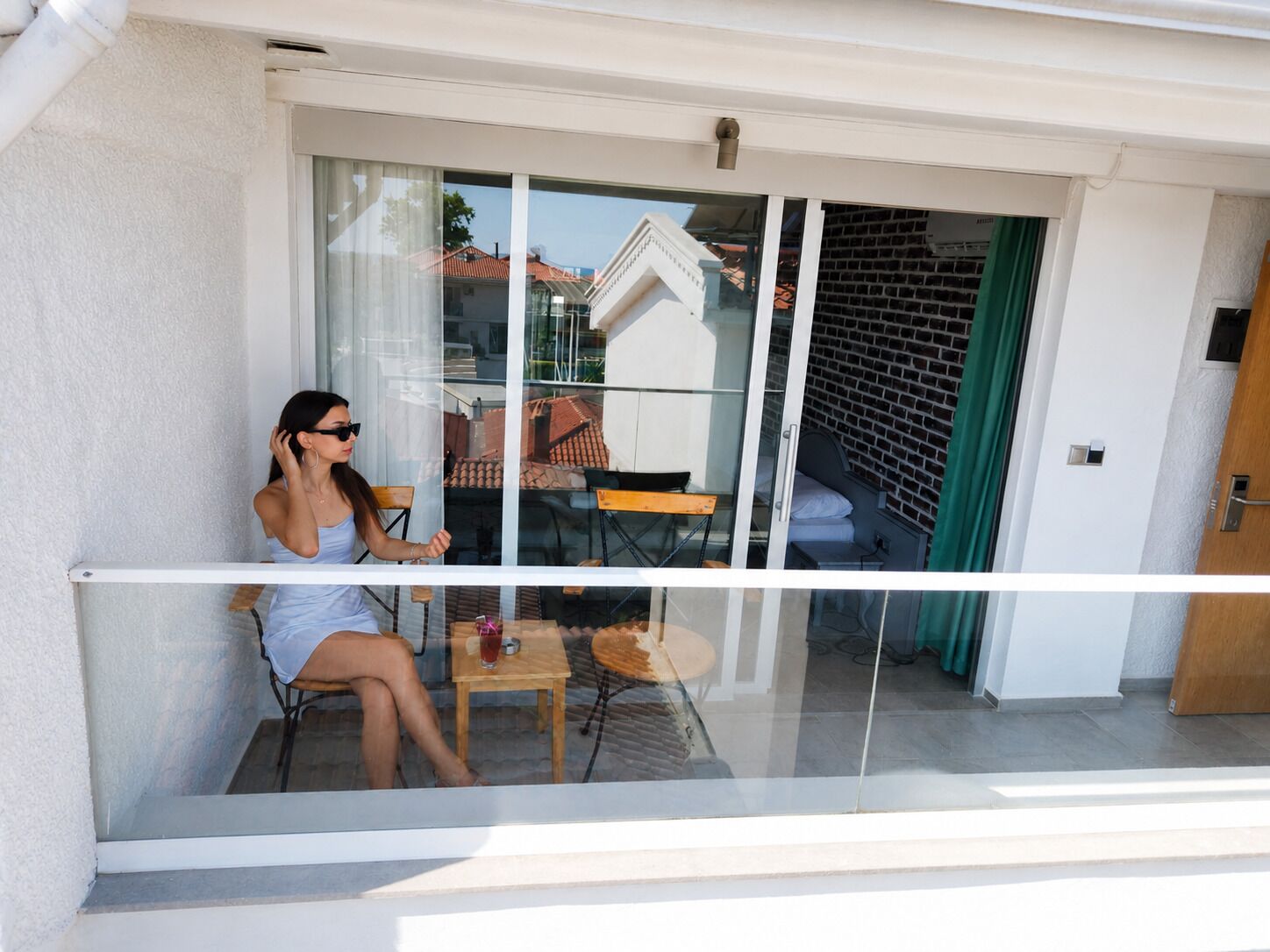 A balcony with a glass door, a chair, a small table, and a view of a building with a brick wall and a window.