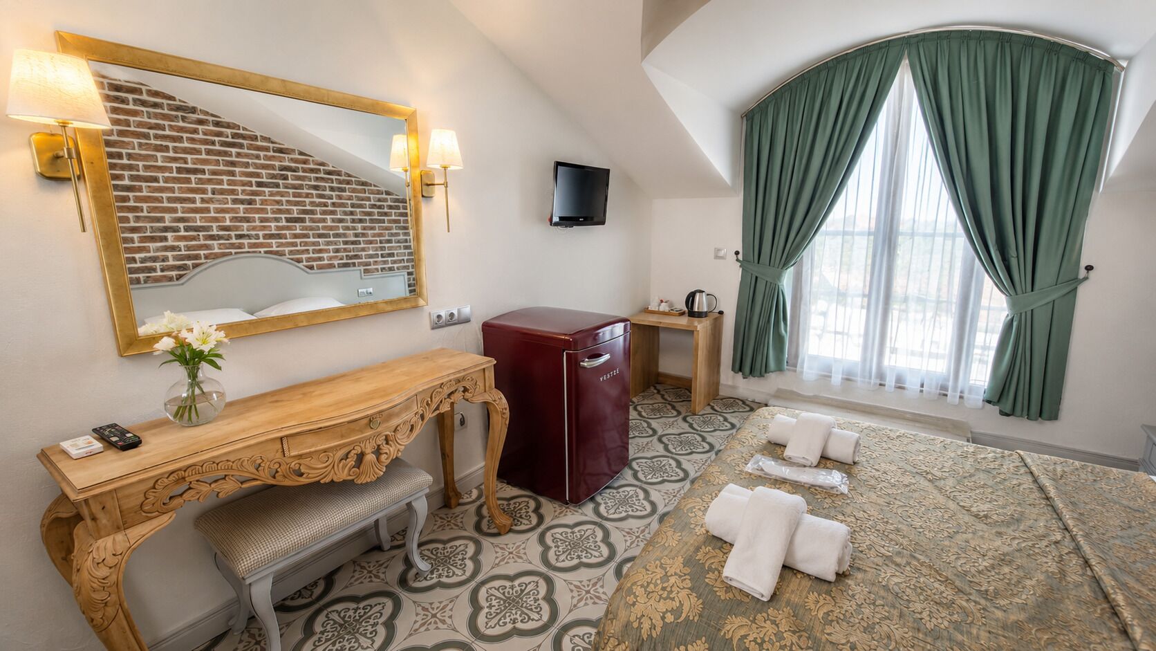 A hotel room with a wooden vanity, a red cabinet, a large mirror, a television, a window with green curtains, and a patterned bedspread.