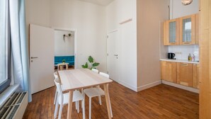 Moderni keittiö, jossa on puiset kaapit, ruokailutila pöytineen ja tuoleineen sekä näkymä avonaisesta ovesta.