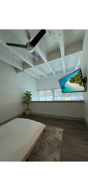 Ein Zimmer mit Deckenventilator, einem flachbildfernseher an der Wand, einer Topfpflanze, einem Bett und einem Fenster mit Blick auf einen Strand.