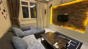 A modern living room with a sectional sofa, a coffee table, a TV mounted on a brick wall, and a window with curtains. - Nova Park Suites (İstanbul)
