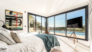Ein Schlafzimmer mit einem großen Bett, einem Fernseher auf einem Schrank und Blick auf Pool und Landschaft durch raumhohe Fenster.