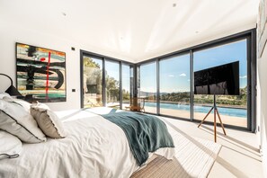 A bedroom with a large bed, a TV on a stand, and a view of a pool and landscape through floor-to-ceiling windows. - Luxury Villa Arizona - Sea View (Grimaud)