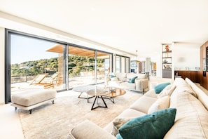 A modern living room with a large glass wall, a sofa set, a coffee table, and a view of a hillside. - Luxury Villa Arizona - Sea View (Grimaud)