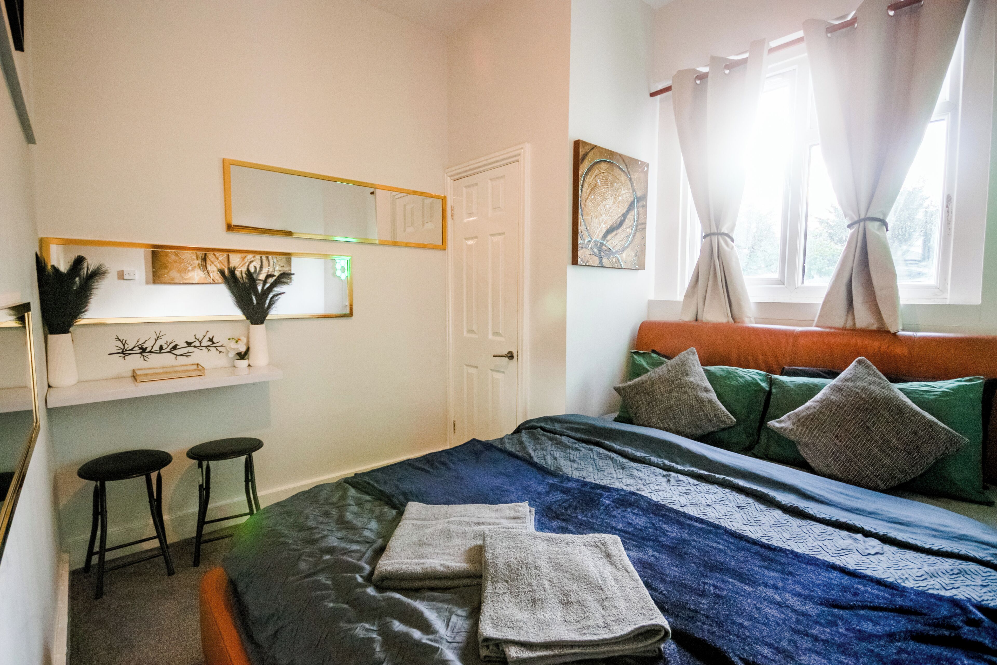 A bedroom with a bed, two stools, a mirror, a door, and a window with curtains.