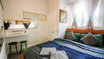 A bedroom with a bed, two stools, a mirror, a door, and a window with curtains.