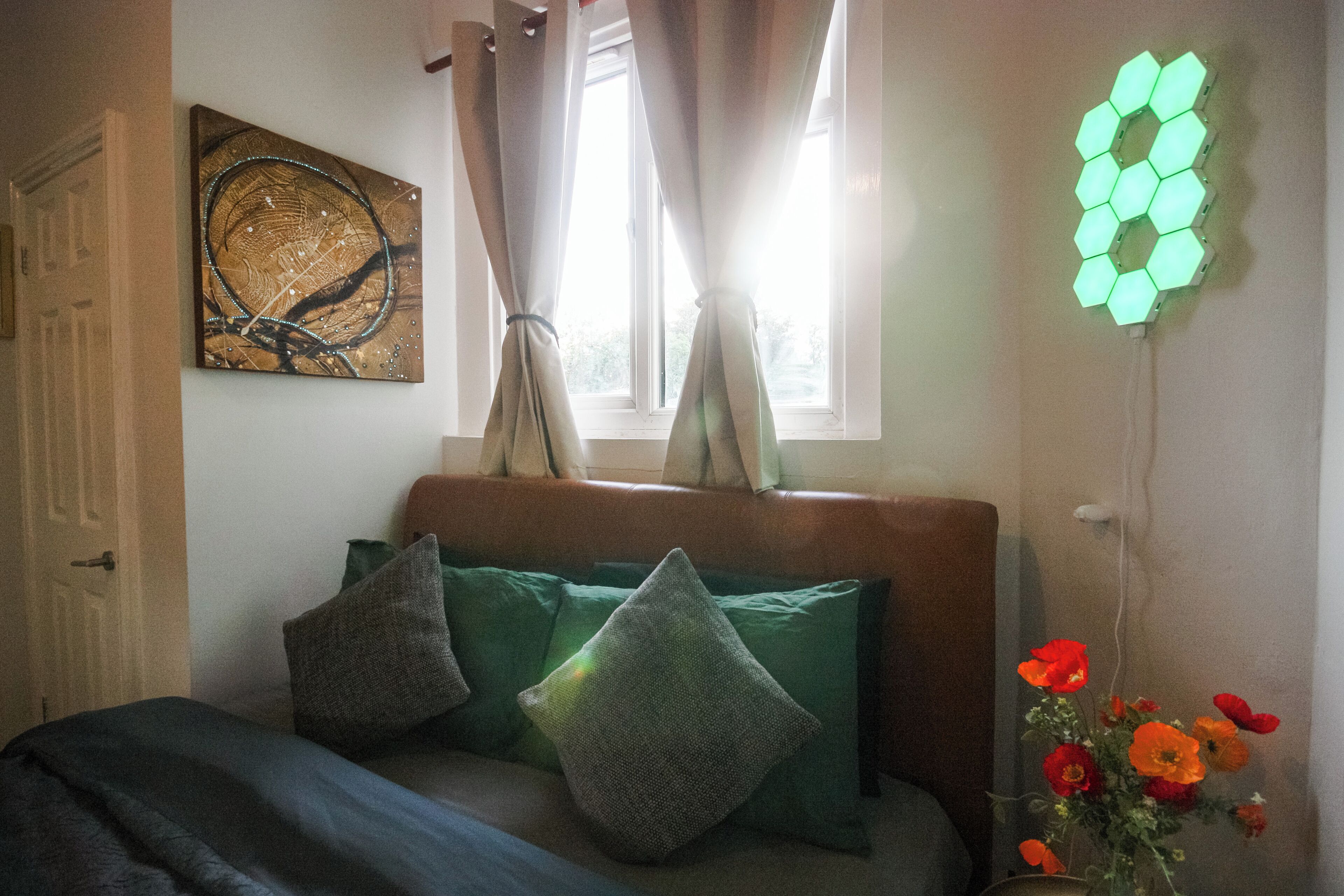 A room with a bed, a window with curtains, a wall-mounted artwork, and a wall light fixture.