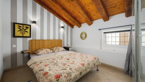 Een slaapkamer met een houten plafond, een bed met bloemenbeddengoed, een wandlamp, een raam met jaloezieën en een gestreept wanddecor.