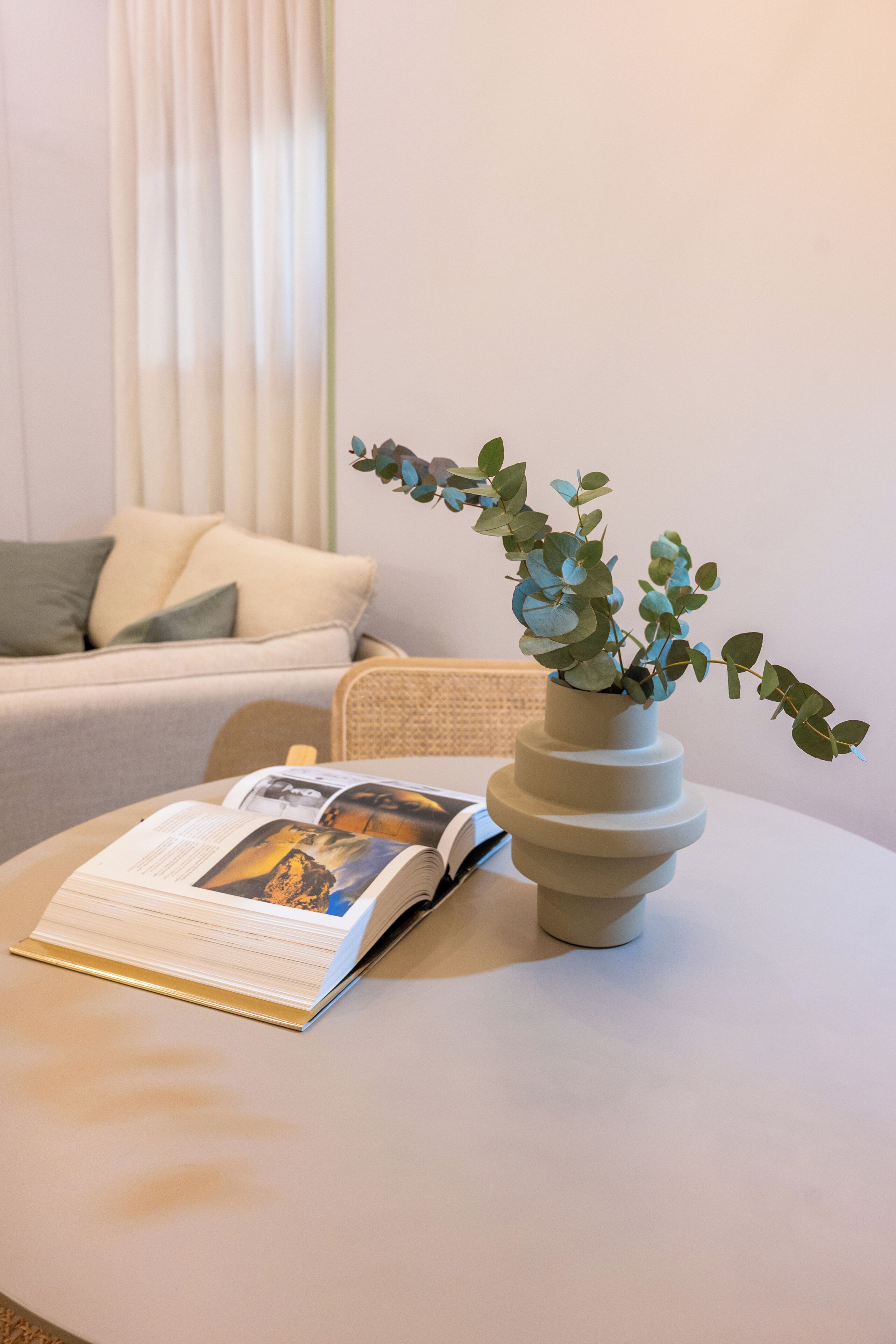 Una habitación con un sofá blanco, una mesita redonda con un libro y un jarrón de hojas verdes.