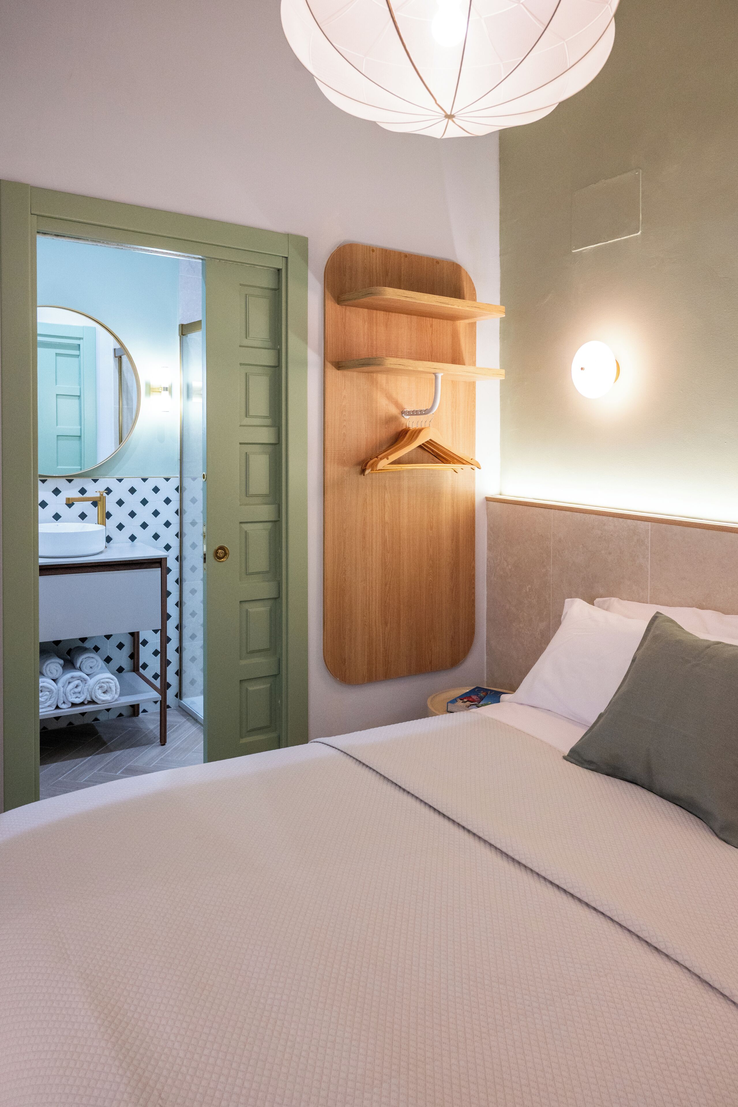 Une chambre à coucher comprenant un lit, une étagère en bois, un porte-vêtements suspendu, une table de chevet et une lampe fixée au mur.