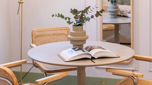 Een kamer met een ronde tafel, rieten stoelen, een potplant, een boek, een spiegel en een vloerlamp.