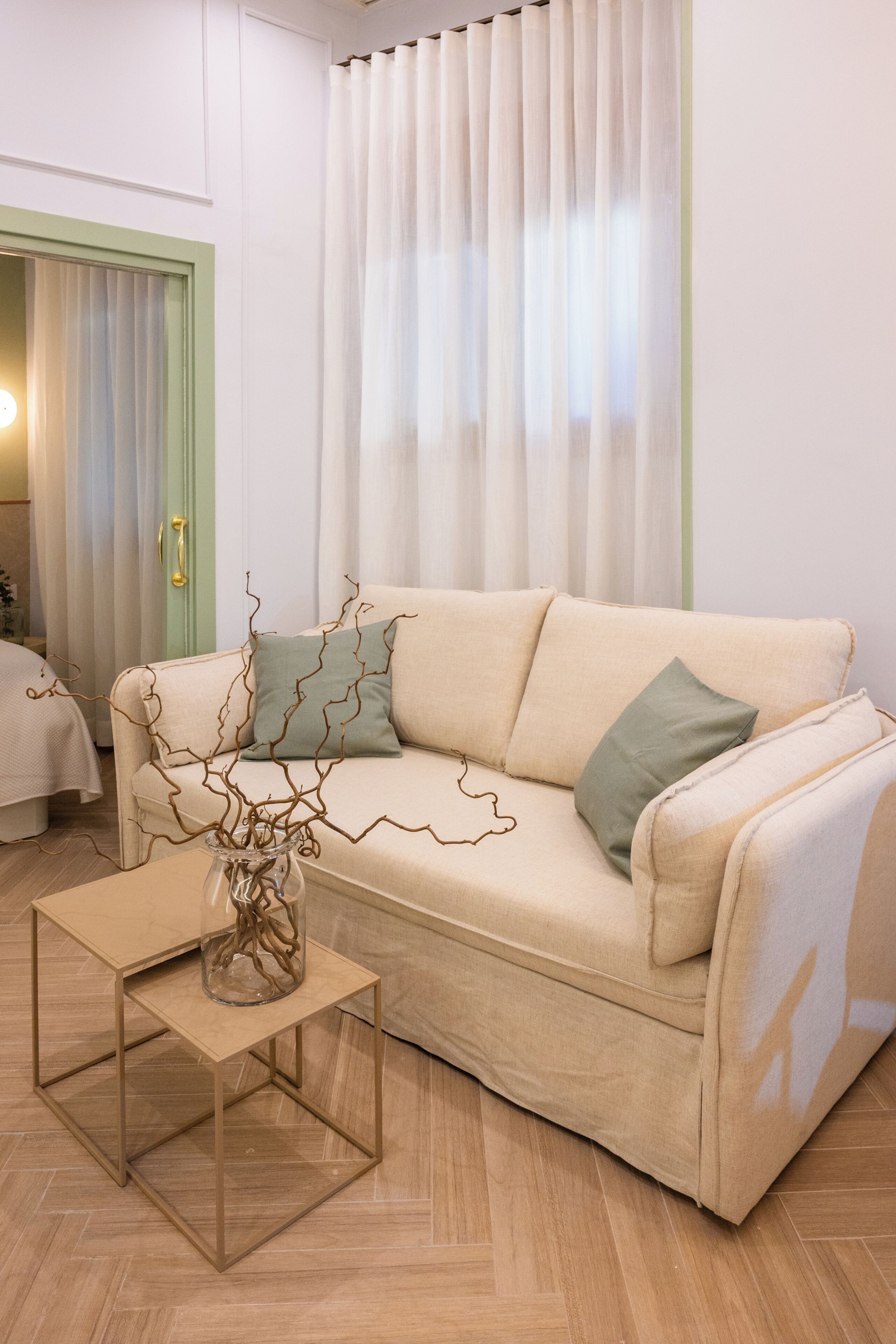 Una sala de estar con un sofá beige, un jarrón de vidrio con ramas, una mesita de madera y una puerta verde.