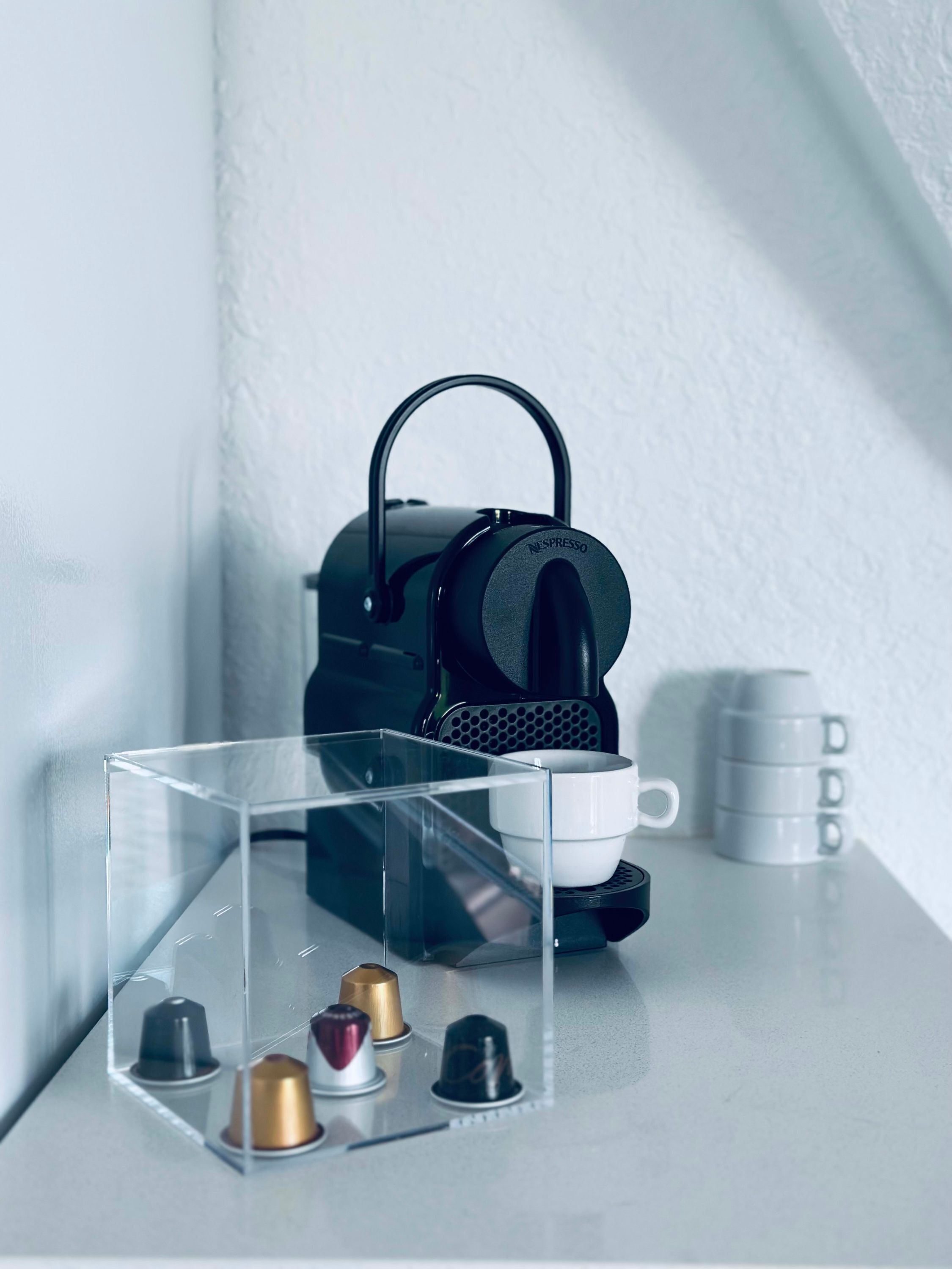 Una máquina de café Nespresso sobre una superficie blanca, con una vitrina transparente que contiene varias cápsulas.