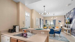 A compact kitchen with a dishwasher, a sink, and a dining area with a blue chair and a table. - Kissimmee 4BR l Resort Living l Great Location (Kissimmee)