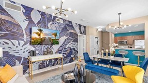 A modern living room with a large wall mural, a dining area with blue chairs, and a kitchen in the background. - Kissimmee 4BR l Resort Living l Great Location (Kissimmee)
