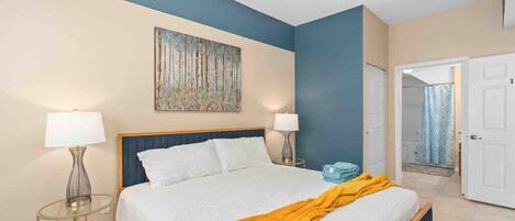 A bedroom with a bed, two bedside lamps, a painting, and a bathroom door.