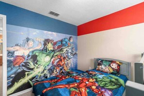 Un dormitorio con un gran mural de superhéroes en la pared, una cama con ropa de cama temática de superhéroes y una pared de acento rojo y blanco.