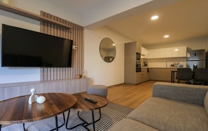 A modern living room with a flat-screen TV, a round wooden coffee table, a grey sofa, and a kitchen in the background. - w Trendy Cozy 2BR w Balcony in Miraflores (Lima)