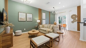 Ein modernes Wohnzimmer mit einer Couch, einem Couchtisch und einem Hocker, ein Essbereich mit Stühlen und eine durch eine offene Tür sichtbare Küche.