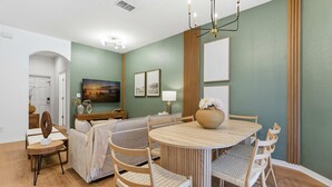 Ein modernes Wohnzimmer mit einem runden Holzesstisch, hellen gewebten Stühlen, einem Sofa und einem Fernseher.