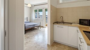 Cocina compacta con lavamanos, microondas y armarios. Se ve una cama en la habitación contigua.