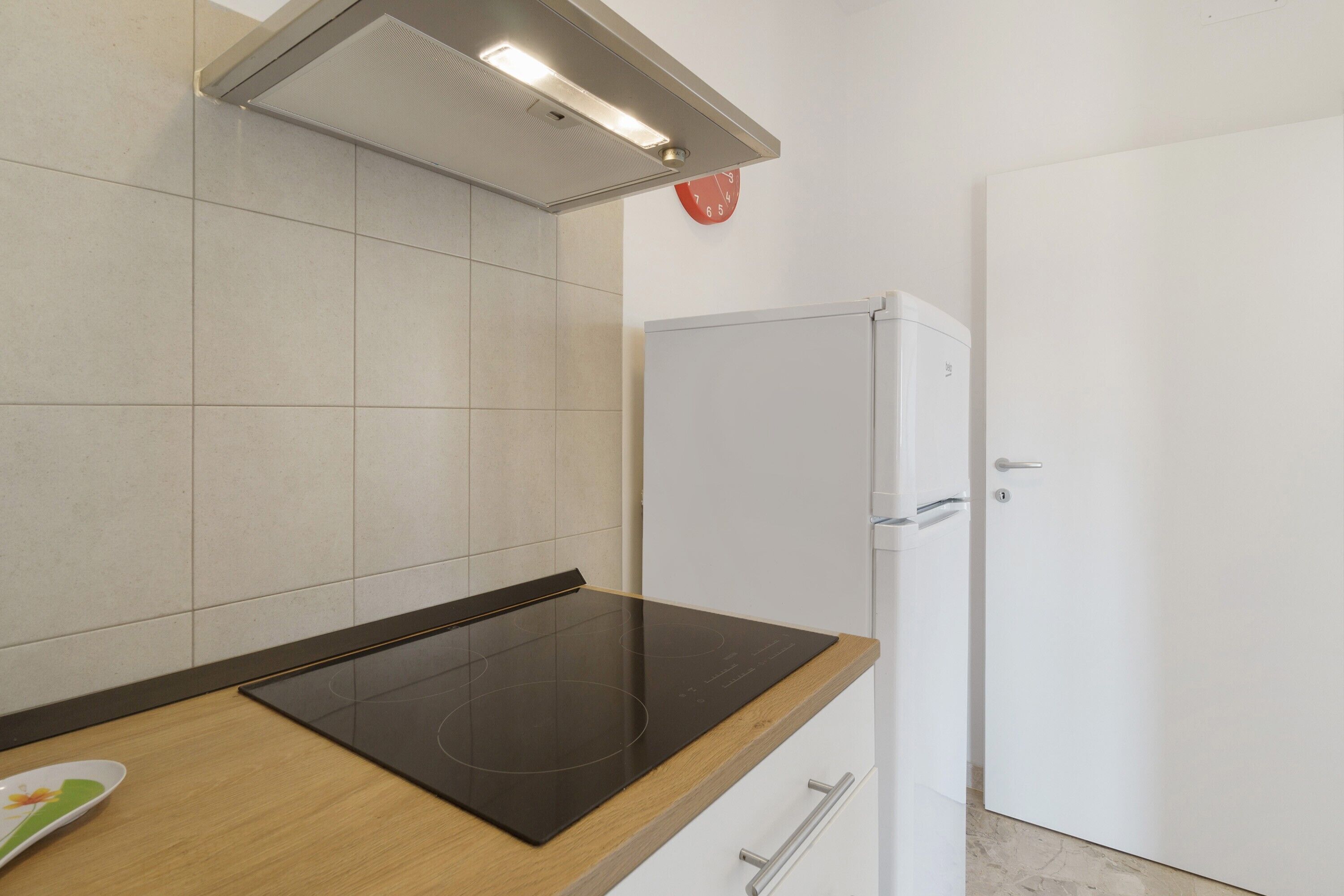 Eine Küche mit einem schwarzen Induktionskochfeld, weißen Schränken, einem weißen Kühlschrank und einer roten Uhr an der Wand.
