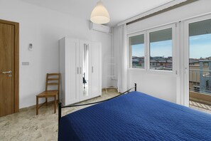 Ein Schlafzimmer mit Bett, Kleiderschrank, Stuhl, einer Pendelleuchte und einem Fenster mit Blick auf Gebäude.
