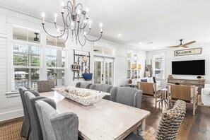 A spacious dining area with a large table, grey upholstered chairs, a chandelier, and a television mounted on the wall.