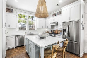 A modern kitchen with white cabinets, a stainless steel refrigerator, a large island with a bowl, and a hanging light fixture.