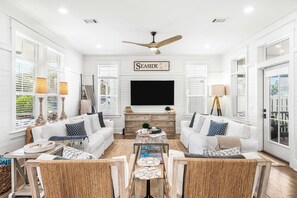 A living room with white walls, a ceiling fan, a flat-screen TV, two white sofas, wicker chairs, a coffee table, and a fireplace.