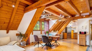 Un loft spacieux avec de hauts plafonds, des poutres apparentes, un coin repas doté de chaises modernes, une cuisine équipée d’appareils électroménagers en acier inoxydable et un coin salon confortable avec un canapé et des coussins.