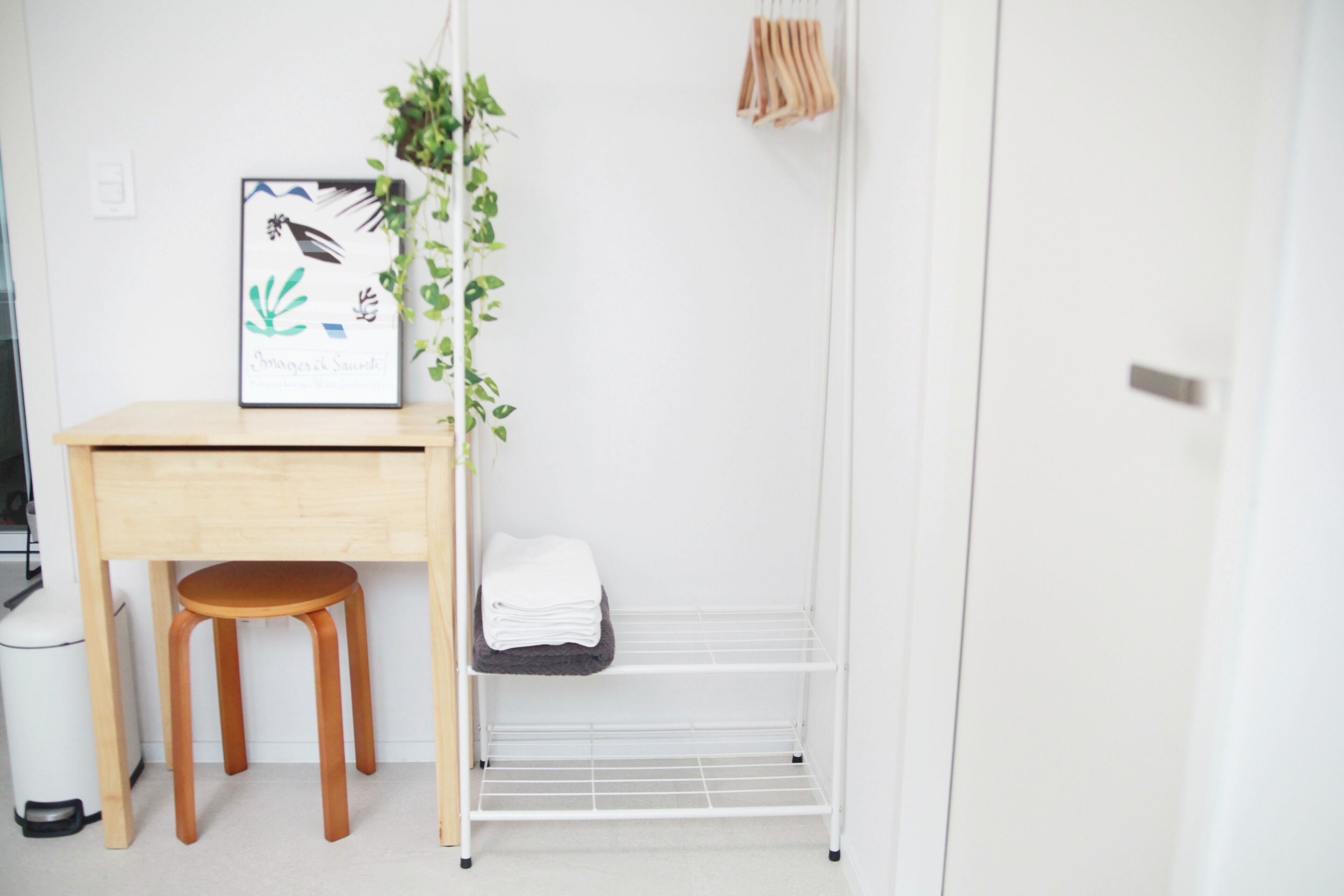 A clean, minimalistic room with a wooden desk, a chair, a white shelf, a framed picture, and a hanging plant.