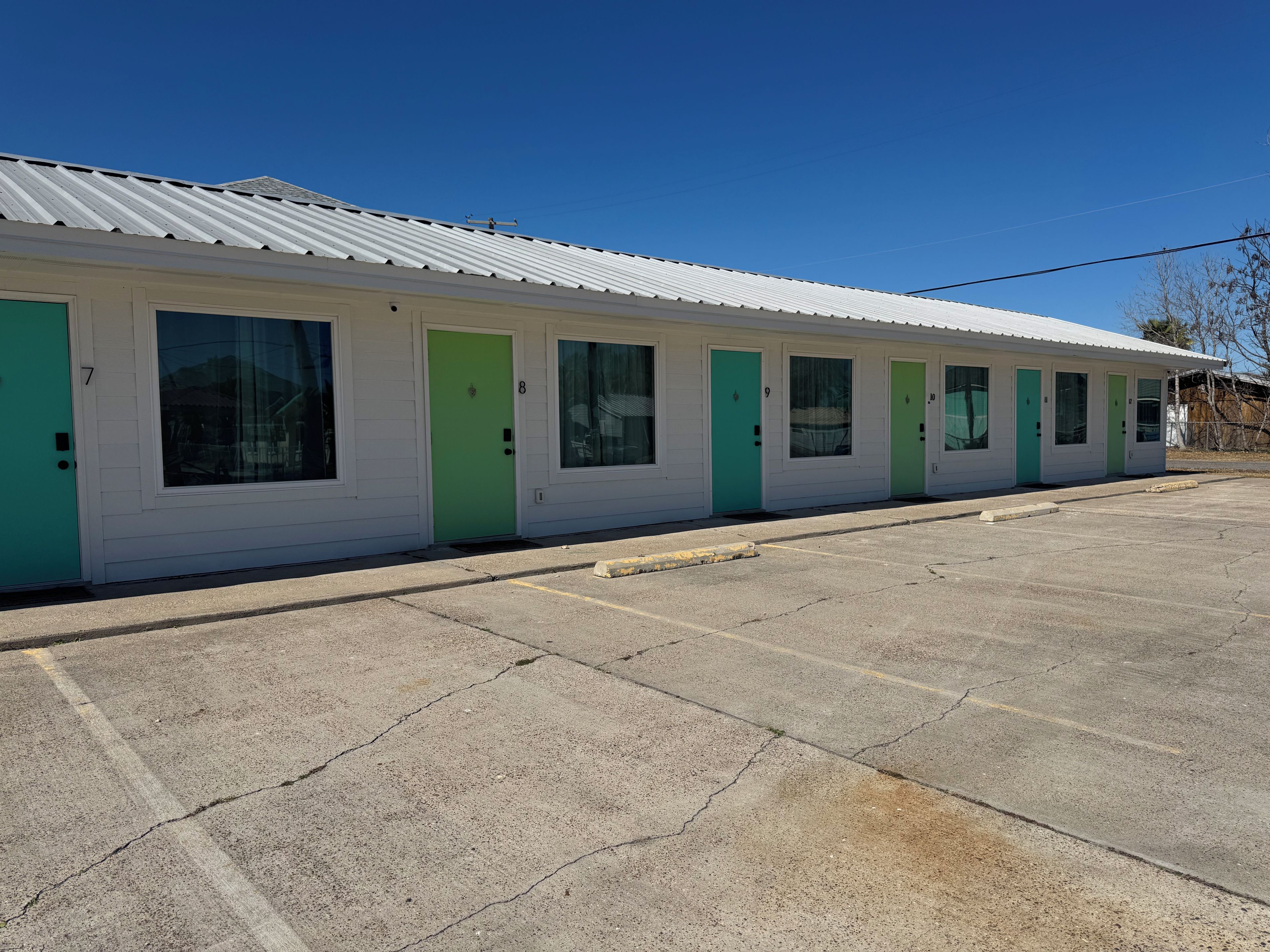 Una hilera de habitaciones de motel con puertas numeradas, un estacionamiento y un cielo azul despejado.