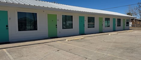 Una hilera de habitaciones de motel con puertas numeradas, un estacionamiento y un cielo azul despejado.