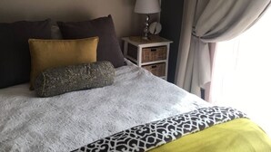 Ein Schlafzimmer mit einem Bett, einem Nachttisch, einer Lampe, zwei gerahmten Bildern an der Wand und einem Fenster mit Vorhängen.