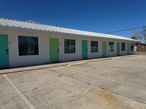 Une rangée de chambres de motel, chacune avec une porte numérotée, un parking et un ciel bleu dégagé.