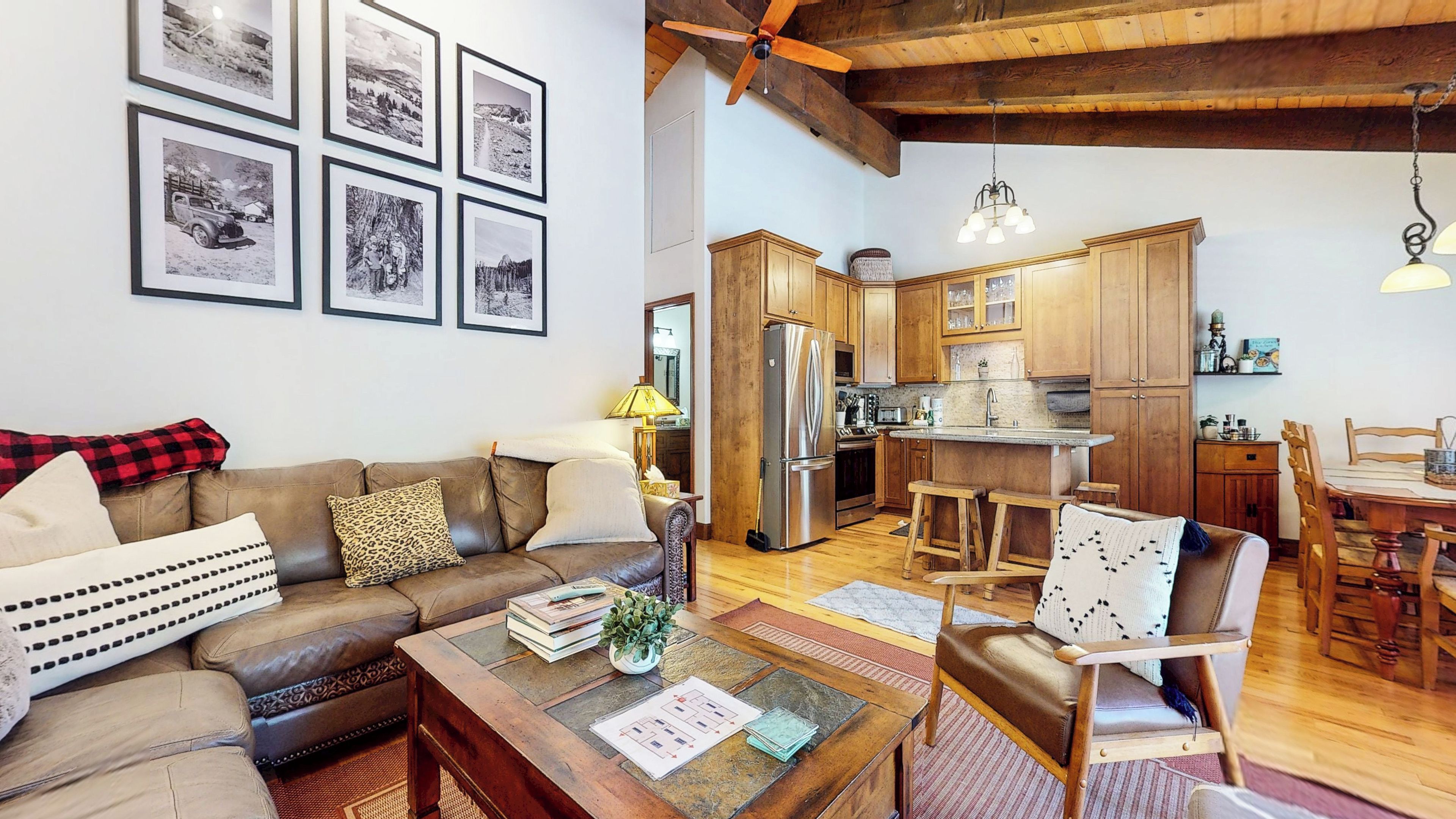 A living room with a sofa, coffee table, and chairs, a kitchen with wooden cabinets and appliances, and a ceiling fan.