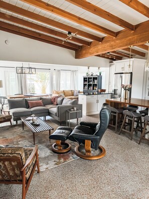 A spacious living area with a high ceiling, a sofa set, a coffee table, a leather chair, and a kitchen area with cabinets and a refrigerator. - Renovated Mid-Century Modern 3 bedroom Home with Fenced In Backyard in Orlando (Orlando)